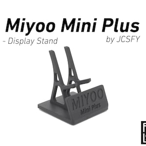 Puede incluir: Un soporte de exhibición negro para el dispositivo de juegos portátil Miyoo Mini Plus. El soporte presenta una base rectangular y una estructura de soporte en ángulo con el texto "MIYOO Mini Plus". Diseño de JCSFY.