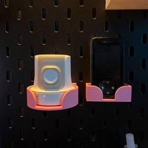 Puede incluir: Dos soportes rosas impresos en 3D, uno para un dispositivo con el texto "dexcom" y otro para un dispositivo negro. Ambos están montados en una panel perforado negro.