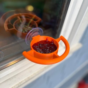 May include: Orange plastic hummingbird feeder with a suction cup attached to a window. The feeder is filled with red liquid.