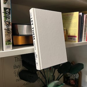 Könnte beinhalten: Ein Hardcover-Buch mit dem Titel "THE LET THEM THEORY" von Mel Robbins steht auf einem Regal. Das Buch hat einen weißen Einband mit schwarzem Text. Weitere Bücher sind sichtbar, darunter eines mit "99 Days" auf dem Buchrücken.