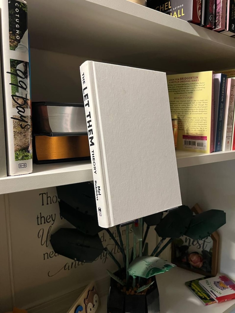 May include: A hardcover book titled "THE LET THEM THEORY" by Mel Robbins stands on a shelf. The book has a white cover with black text. Other books are visible on the shelf, including one with "99 Days" on the spine.