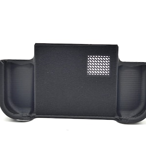 May include: A black, rectangular device with two curved side compartments and a central section. The central section has a square, perforated vent. The device is made of a matte material and appears to be 3D-printed.