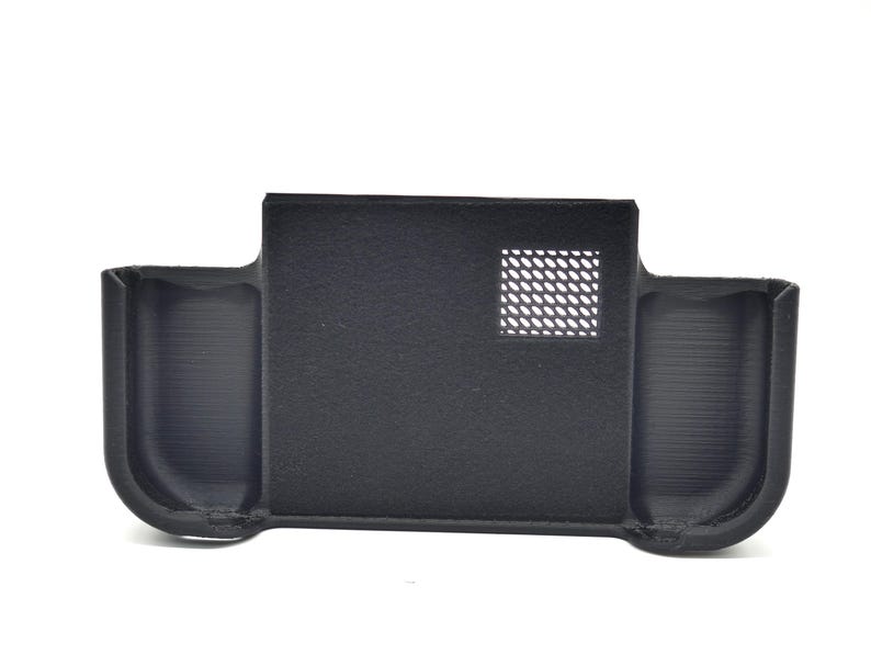 May include: A black, rectangular device with two curved side compartments and a central section. The central section has a square, perforated vent. The device is made of a matte material and appears to be 3D-printed.