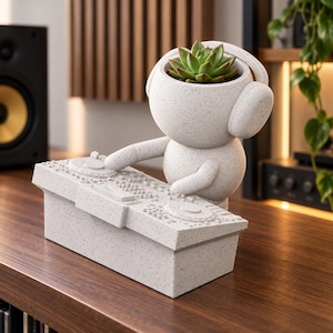 Pode incluir: Um vaso cinzento-claro salpicado em forma de personagem com auscultadores a operar uma mesa de mistura de DJ. A cabeça da personagem serve de recipiente para uma planta suculenta verde, com a figura posicionada atrás de uma consola retangular.