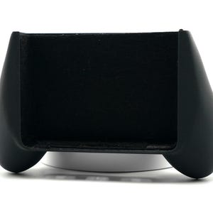 May include: A black gaming controller grip with a curved design. The grip has a rectangular slot in the center and rounded handles on each side. The controller is designed to enhance the gaming experience.