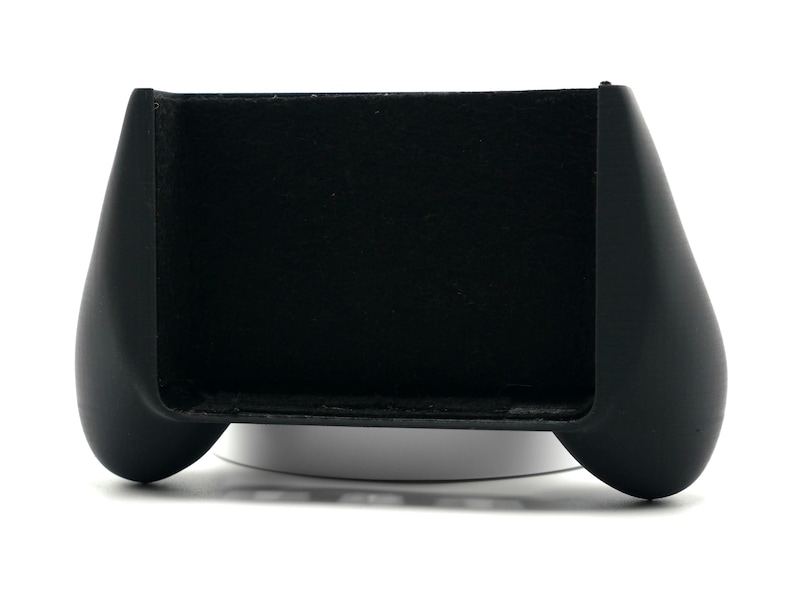 May include: A black gaming controller grip with a curved design. The grip has a rectangular slot in the center and rounded handles on each side. The controller is designed to enhance the gaming experience.
