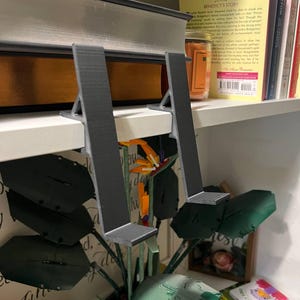 May include: A pair of gray, 3D-printed bookends supporting a large book on a white shelf. The bookends have a simple, angular design. A decorative plant with large green leaves and a colorful flower is in front of the shelf.
