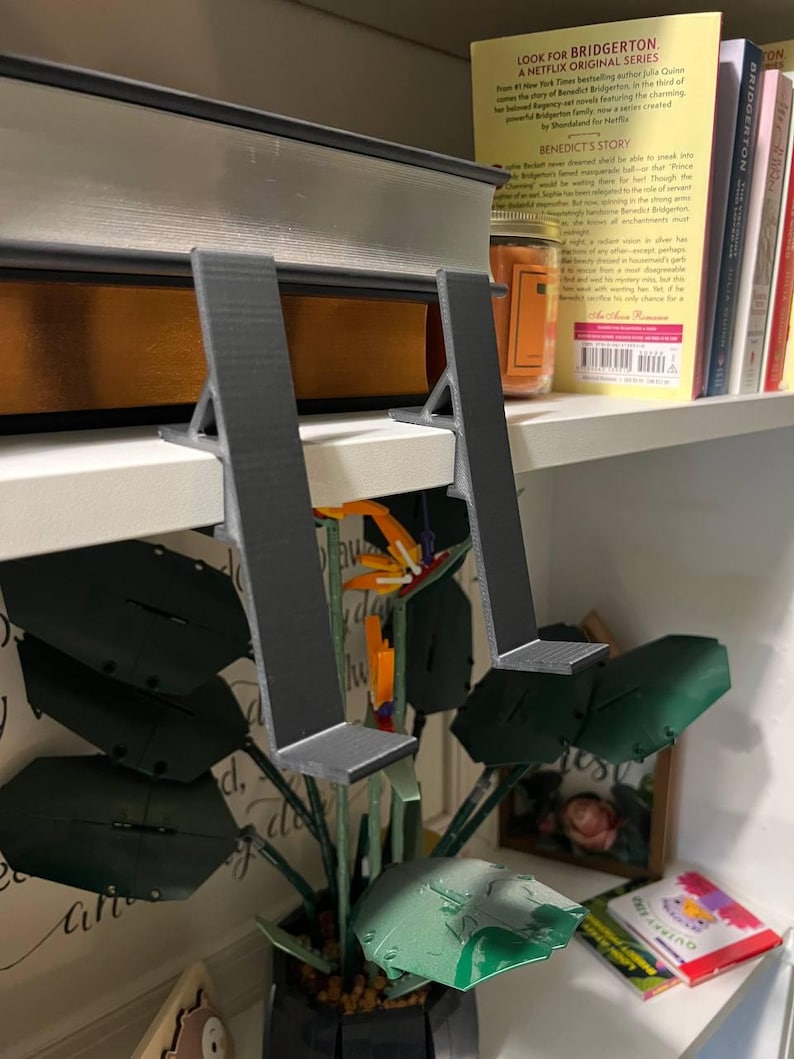 May include: A pair of gray, 3D-printed bookends supporting a large book on a white shelf. The bookends have a simple, angular design. A decorative plant with large green leaves and a colorful flower is in front of the shelf.