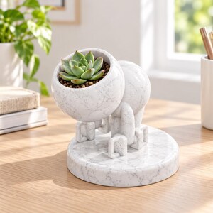 May include: A white marble-patterned planter shaped like a small robot with four square feet and a round head. The head features a hollow opening containing a green succulent plant. The entire structure rests on a circular marble-patterned base.