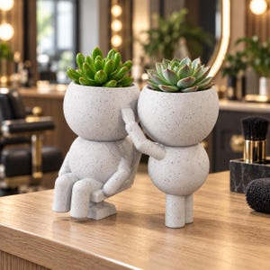 Puede incluir: Dos macetas de cerámica gris claro con motas en forma de figuras estilizadas. Una figura está sentada en una silla pequeña mientras la otra está de pie detrás, pareciendo peinar su cabello. Cada cabeza contiene una planta suculenta verde.