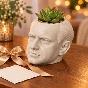 May include: A light gray ceramic planter shaped like a human head with a realistic facial structure, including detailed eyes, nose, and ears. A green succulent plant is potted in the top of the head, surrounded by small brown pebbles.