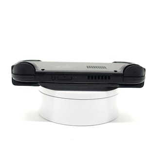 May include: A black handheld gaming console with rounded edges, resting on a white rotating platform. The console features buttons, vents, and a speaker. The device is designed for portable gaming.