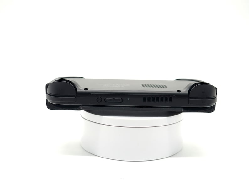 May include: A black handheld gaming console with rounded edges, resting on a white rotating platform. The console features buttons, vents, and a speaker. The device is designed for portable gaming.