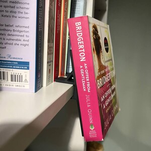 May include: A pink hardcover book titled "Bridgerton: An Offer From a Gentleman" by Julia Quinn, displayed on a white shelf. The book has a woman's silhouette on the cover and is propped up by a black book stand.