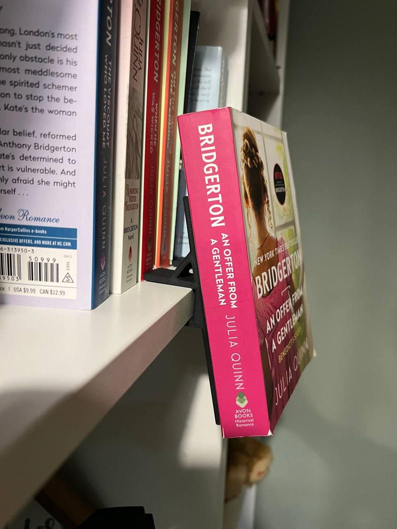 May include: A pink hardcover book titled "Bridgerton: An Offer From a Gentleman" by Julia Quinn, displayed on a white shelf. The book has a woman's silhouette on the cover and is propped up by a black book stand.