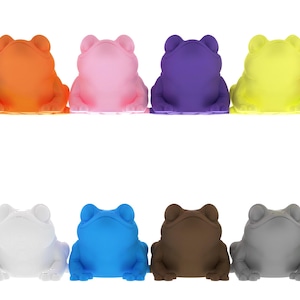 May include: A collection of twelve frog figurines in various colors, including blue, orange, pink, purple, yellow, red, black, white, blue, brown, gray, and green. The frogs are arranged in two rows, each with six frogs.