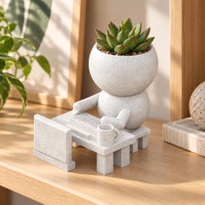 May include: A white speckled ceramic planter shaped like a figure sitting at a desk with a computer monitor, keyboard, and mug. The head of the figure is a round pot containing a green succulent plant, while the body rests on a matching desk.