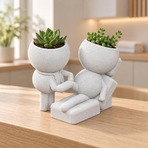 May include: A speckled white ceramic planter set featuring two stylized figures. One figure stands as a doctor with a stethoscope, while the other reclines on an examination table. Each figure has a round head serving as a pot for green succulents.