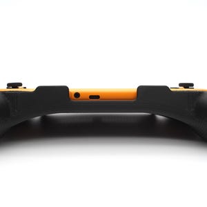 Op de afbeelding: Een zwarte en oranje game controller grip. De grip heeft een gebogen ontwerp met getextureerde handgrepen voor meer comfort. De controller heeft twee zwarte knoppen aan de bovenkant en een oranje middengedeelte. Het ontwerp is strak en modern.