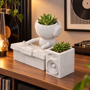 Pode incluir: Um vaso branco salpicado em forma de DJ com auscultadores, posicionado atrás de um vaso retangular de consola de DJ e um vaso quadrado em forma de altifalante. Cada vaso contém uma pequena planta suculenta verde, criando um conjunto.
