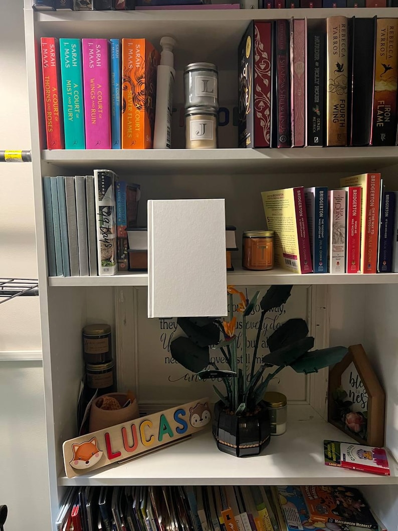 May include: A white bookshelf filled with books of various colors and sizes. Decorative items include candles, a wooden name puzzle that says "LUCAS", and a small potted plant. The books are the main focus, suggesting a literary theme.