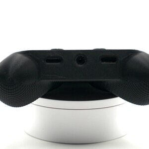 May include: A black gaming controller accessory with textured grips. The controller has a central panel with ports and a rounded shape for comfortable handling. It is resting on a white, circular stand.