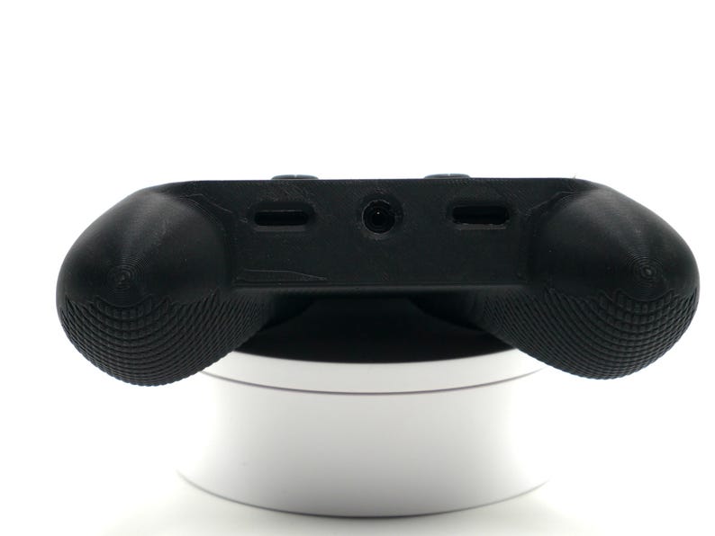 May include: A black gaming controller accessory with textured grips. The controller has a central panel with ports and a rounded shape for comfortable handling. It is resting on a white, circular stand.