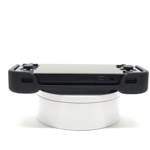 May include: A black handheld gaming console with a protective black rubber case. The console features a screen, buttons, and ports. It is resting on a white rotating display stand. The image is taken against a white background.