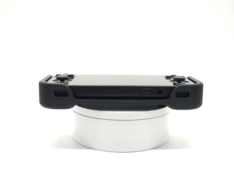 May include: A black handheld gaming console with a protective black rubber case. The console features a screen, buttons, and ports. It is resting on a white rotating display stand. The image is taken against a white background.