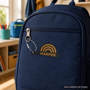 May include: A dark navy blue backpack featuring a black keychain attached to the front zipper. The keychain has a gold-colored rainbow graphic above the name HARPER printed in gold capital letters on a black, elongated, rounded base.