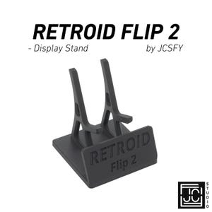 Könnte beinhalten: Ein schwarzer Display-Ständer für die Handheld-Spielkonsole Retroid Flip 2. Der Ständer trägt den weißen Schriftzug "RETROID FLIP 2" und "- Display Stand". Er dient dazu, die Konsole aufrecht zu halten.