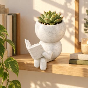 May include: A white speckled ceramic planter shaped like a seated figure reading an open book. The head of the figure serves as a container for a green succulent plant. The figure has a rounded body, arms, and legs, all featuring a consistent speckled texture.