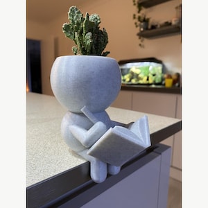 May include: A light gray speckled planter shaped like a stylized figure sitting on an edge while reading an open book. The head of the figure serves as a pot containing a green cactus. The object has a matte, textured finish throughout its form.