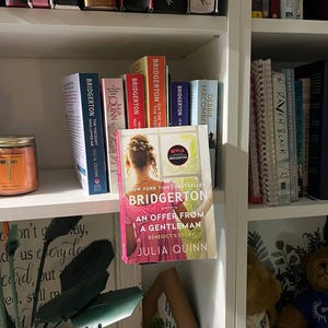 May include: A bookshelf filled with books, including several copies of the "Bridgerton" series by Julia Quinn. The featured book is "An Offer From a Gentleman." The books are arranged on white shelves, with a decorative candle and other items visible.