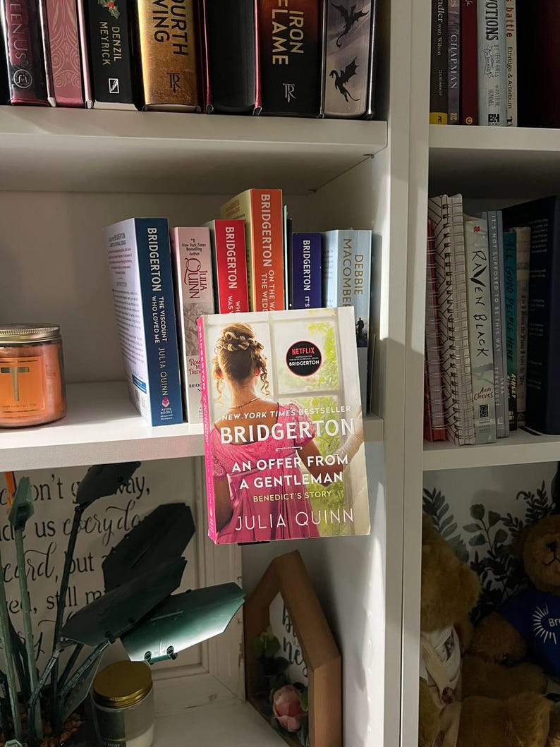 May include: A bookshelf filled with books, including several copies of the "Bridgerton" series by Julia Quinn. The featured book is "An Offer From a Gentleman." The books are arranged on white shelves, with a decorative candle and other items visible.