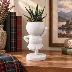 Peut inclure: Un pot de fleurs blanc moucheté en forme de personnage en pose de ballet, avec une tête arrondie, un corsage à petits boutons et une jupe plissée. Le pot contient une plante succulente verte aux feuilles pointues rayées de blanc.