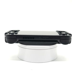 May include: A black handheld gaming console with buttons and a screen, resting on a white rotating stand. The console has a sleek design with a matte finish, and is designed for portable gaming.