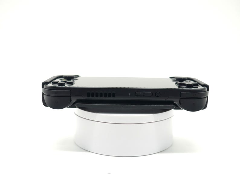 May include: A black handheld gaming console with buttons and a screen, resting on a white rotating stand. The console has a sleek design with a matte finish, and is designed for portable gaming.
