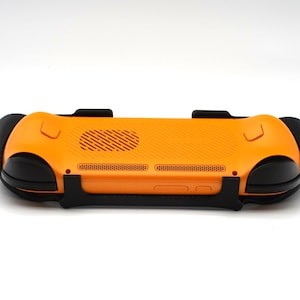 Op de afbeelding: Een oranje en zwarte gameconsole grip, gezien vanaf de achterkant. De grip heeft een getextureerd oranje oppervlak met zwarte handgrepen en een zwarte ondersteuningsstructuur. Het ontwerp bevat ventilatie- en knopdetails.