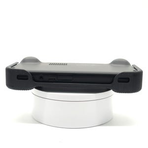 May include: A black handheld gaming device with a protective case. The case has rounded grips on each side and a series of vents along the top. The device is resting on a white, circular stand.