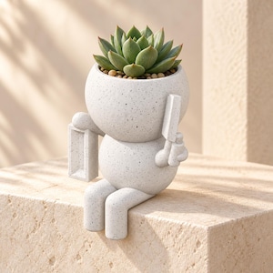 May include: A light gray speckled ceramic planter shaped like a seated figure with a round head and body. The figure holds a rectangular frame in one hand and a small tool in the other. A green succulent plant is potted inside the head section.