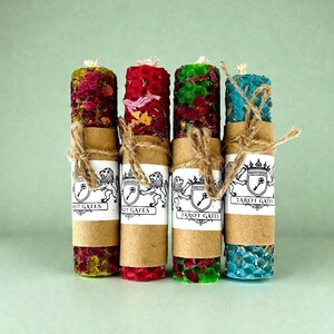 Puede incluir: Cuatro velas de pilar decorativas en tonos rojo, verde y azul, adornadas con flores secas. Cada vela está envuelta con una banda de papel marrón y atada con hilo. La etiqueta en cada vela dice "TAROT GATES".