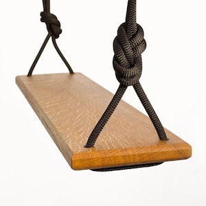 Op de afbeelding: Een houten schommel met een natuurlijke houten afwerking en een zwart touw. De schommel is aan het plafond gehangen en heeft een rechthoekige houten zitting.