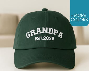 May include: A dark green baseball cap with the word "GRANDPA" embroidered in white, along with "EST.2026". The cap has a curved brim and a button on top. The background is a light color.