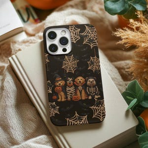 May include: Black phone case featuring a Halloween design with cobwebs, bats, and costumed dogs. The dogs are dressed as a witch, mummy, vampire, and skeleton, each holding a pumpkin. The case is on a book.