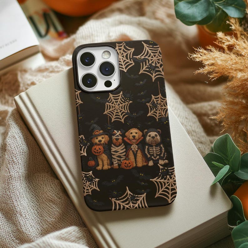 May include: Black phone case featuring a Halloween design with cobwebs, bats, and costumed dogs. The dogs are dressed as a witch, mummy, vampire, and skeleton, each holding a pumpkin. The case is on a book.
