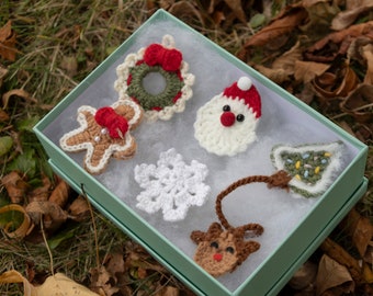 Puede incluir: Una caja de regalo verde con un interior blanco contiene seis adornos navideños de ganchillo: un hombre de jengibre, una corona, un Papá Noel, un copo de nieve, un árbol de Navidad y un reno.