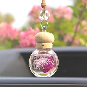 May include: A clear, round glass car air freshener with a wooden top and a decorative crystal bead. The air freshener contains a pink and white flower. It hangs from a brown cord.