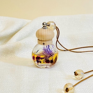 May include: A small, clear glass diffuser with a wooden cap and a crystal bead. The diffuser contains dried purple flowers and a yellow liquid. A brown cord is attached for hanging.