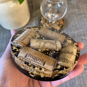 May include: A round black resin coaster with gold leaf accents, embedded with several wine corks. The corks have various winery names and logos printed on them. A wine glass sits nearby.